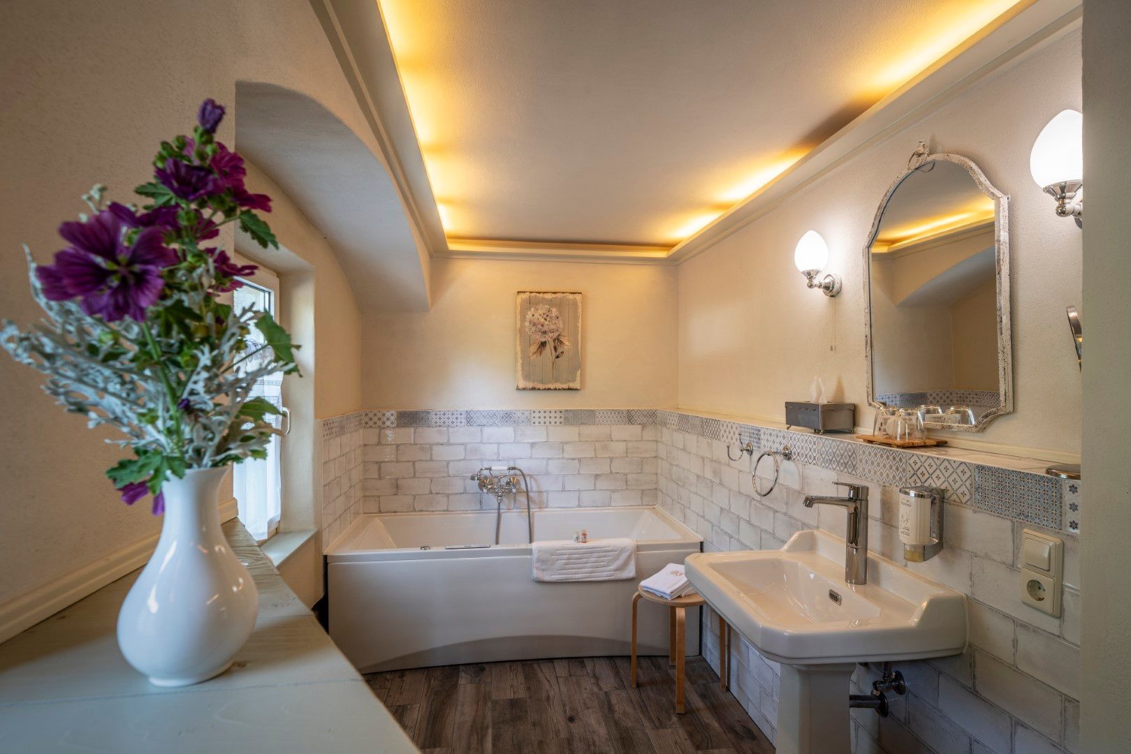 Badezimmer mit weißer Badewanne, Waschbecken und dekorativem Blumenarrangement. Deckenbeleuchtung, weiße Wände und Holzboden.