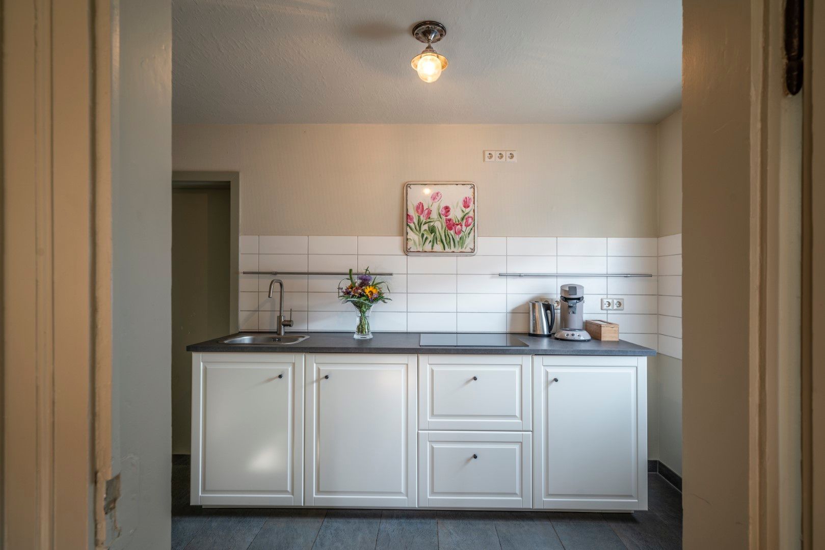 Küche mit weißen Schränken, grauer Arbeitsplatte, Spüle, Küchengeräten und floralen Bildern an einer cremefarbenen Wand.