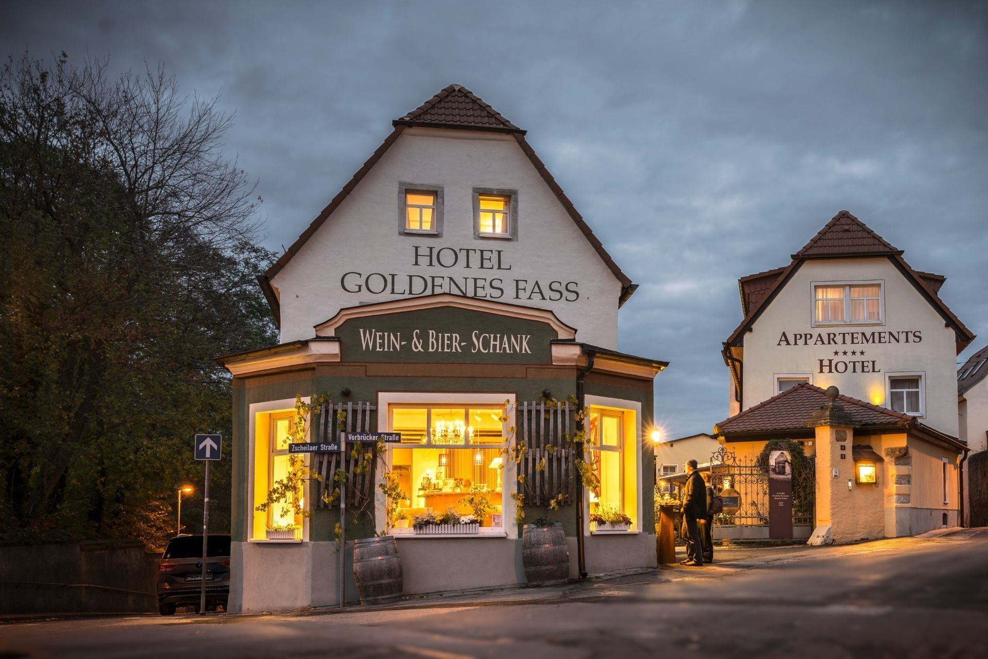 Hotel „Goldenes Fass“ in der Abenddämmerung. Zwei Gebäude mit beleuchteten Fenstern, eines mit der Aufschrift „Wein-Stube“. Eine Person steht am Eingang.