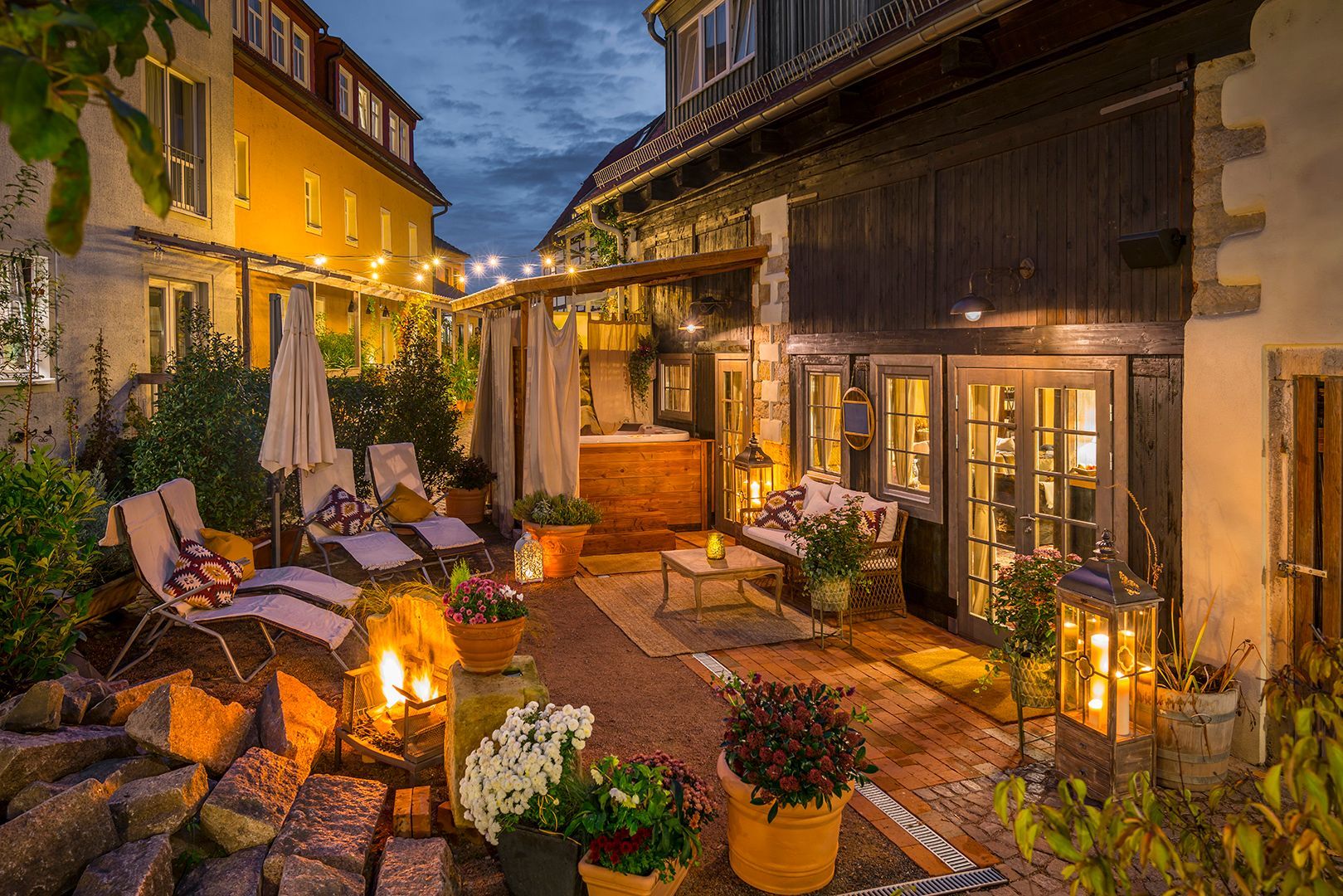 Gemütlicher Innenhof im Freien in der Abenddämmerung mit Sitzgelegenheiten, Laternen und Lichterketten, umgeben von Gebäuden.