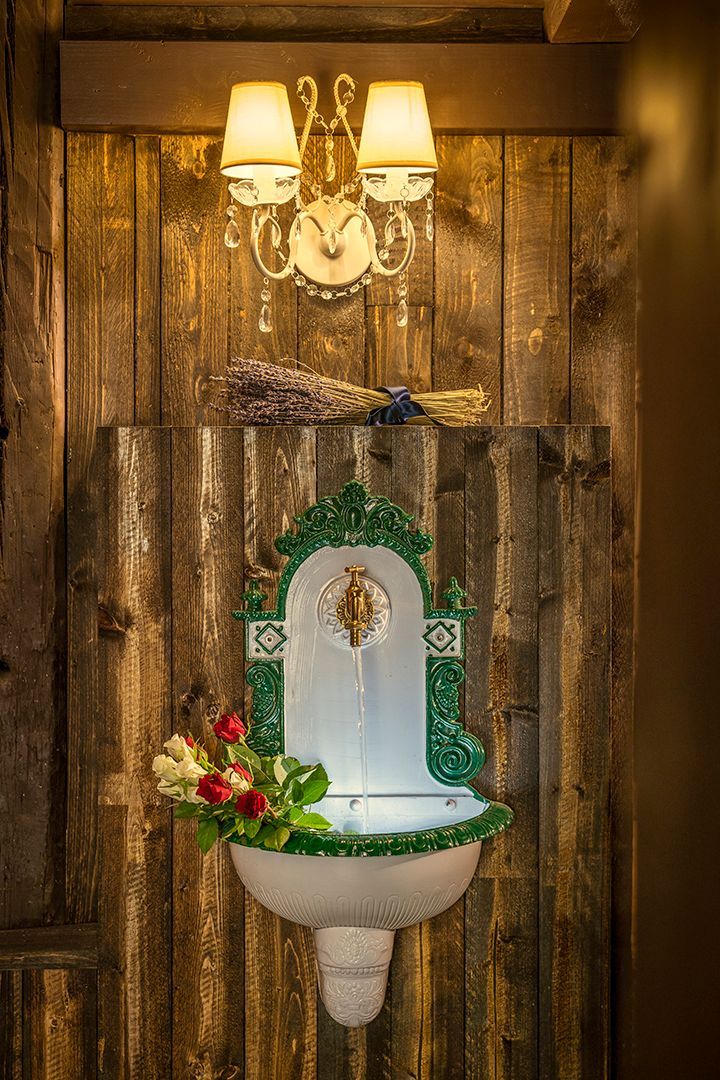 Ein weißer und grüner dekorativer Springbrunnen an einer rustikalen Holzwand, beleuchtet von einem Kronleuchter.