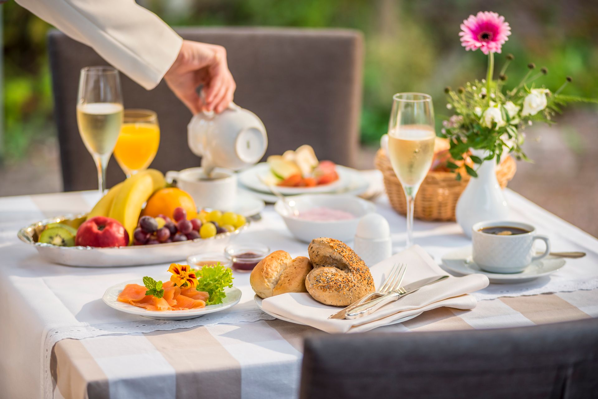 Eine Hand gießt Kaffee ein an einem sonnigen Frühstückstisch im Freien, Obst, Gebäck, Räucherlachs und Champagner.