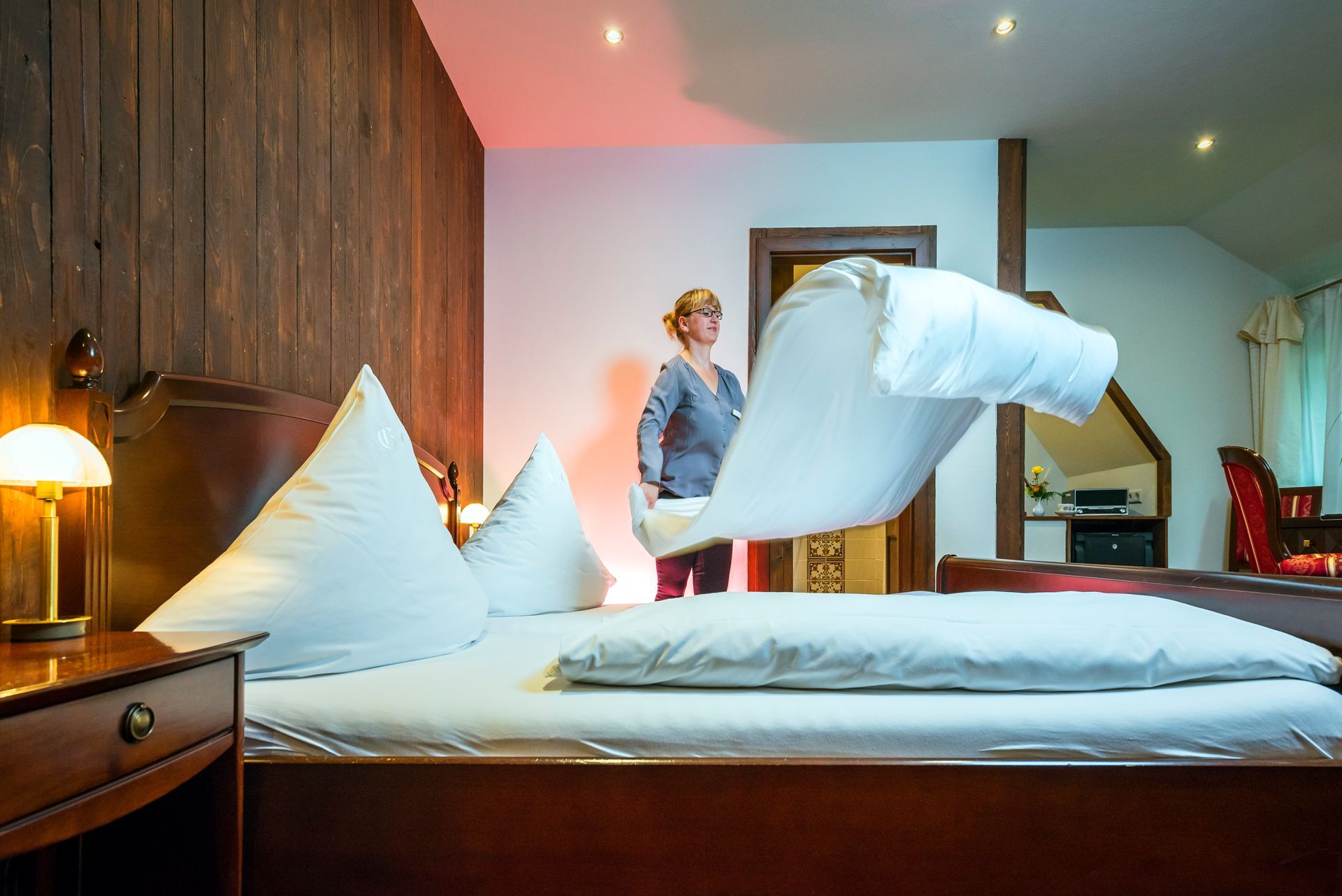 Eine Frau macht in einem Hotelzimmer mit dunkler Holzvertäfelung das Bett und hält dabei eine weiße Bettdecke hoch.