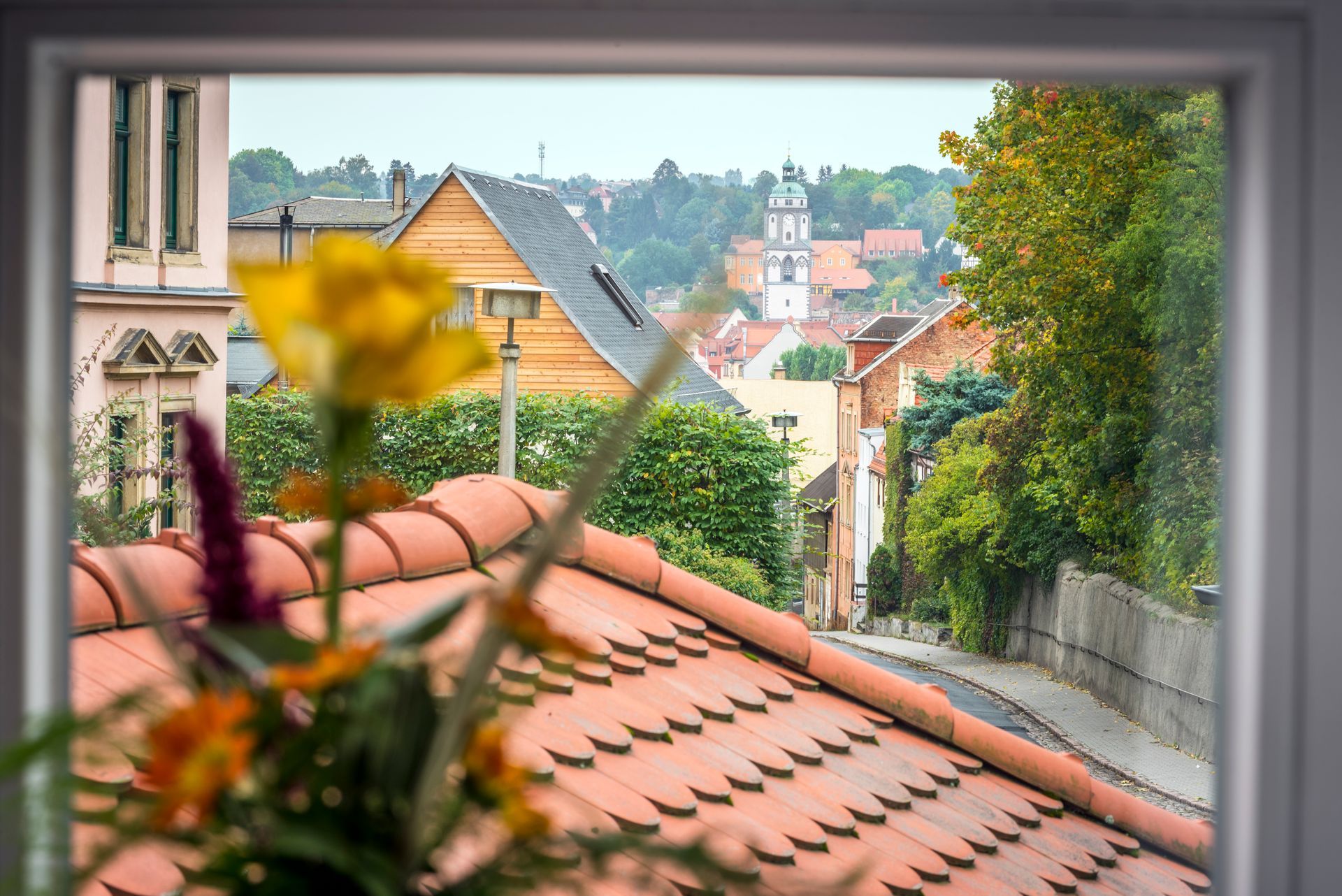 Blick aus einem Fenster auf eine Straße mit roten Ziegeldächern und einem Kirchturm.