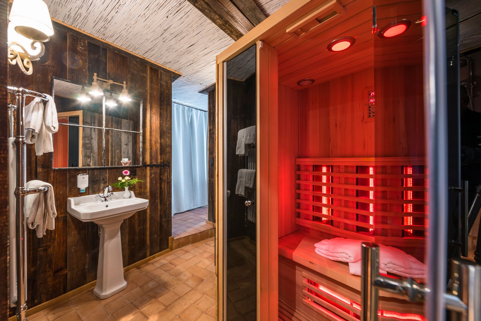 Badezimmer mit Sauna, Holzwänden. Sichtbarer roter Infrarotsauna-Innenraum. Waschbecken, Spiegel, Handtücher.