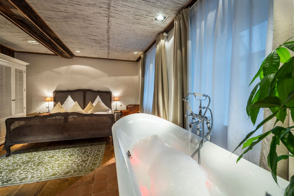 Schlafzimmer mit freistehender Badewanne auf Füßen, großem Bett und Fenster mit Vorhängen. Sanfte Beleuchtung.