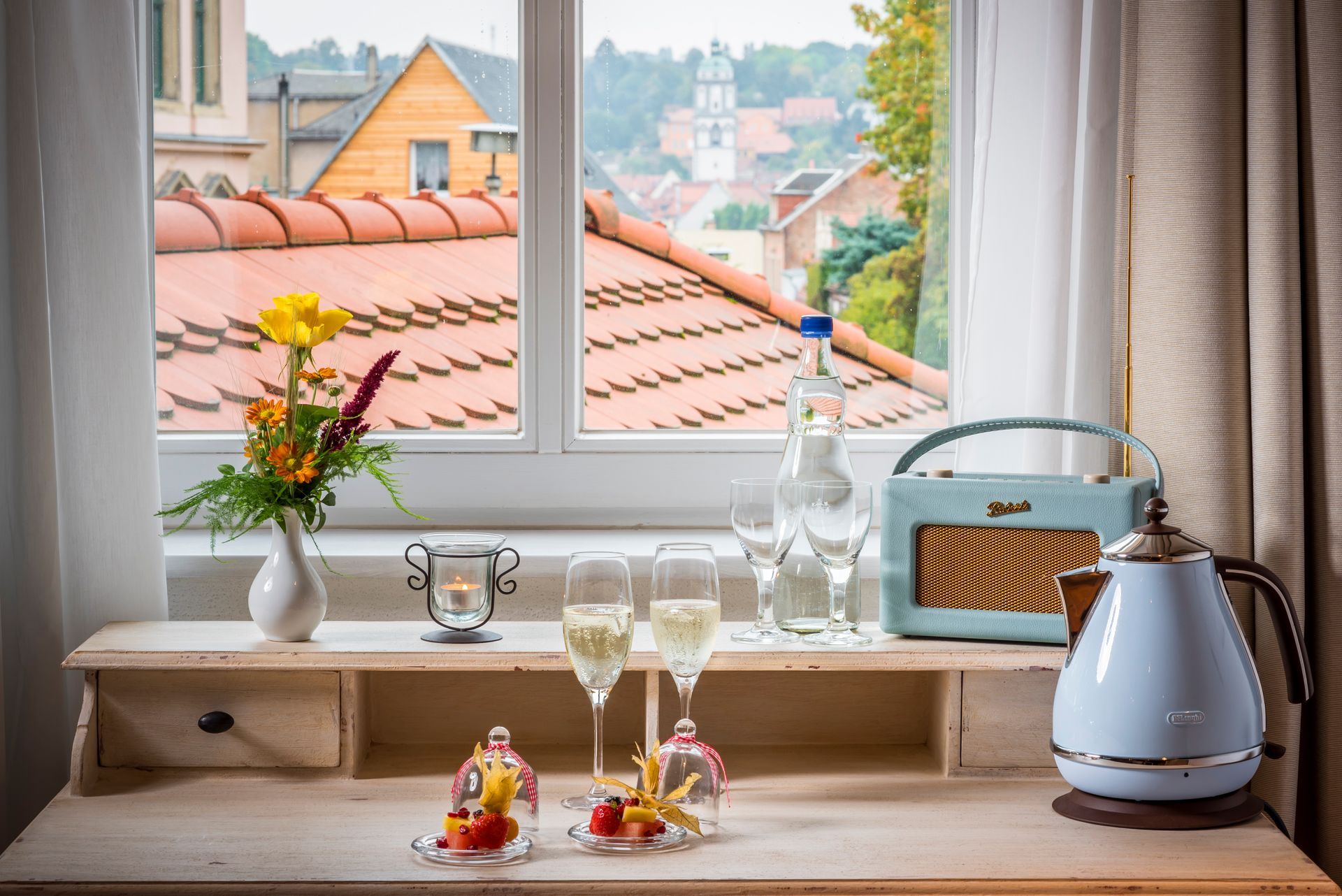 Schreibtisch mit Getränken, Blumen und Wasserkocher an einem Fenster mit Blick auf eine Stadt.
