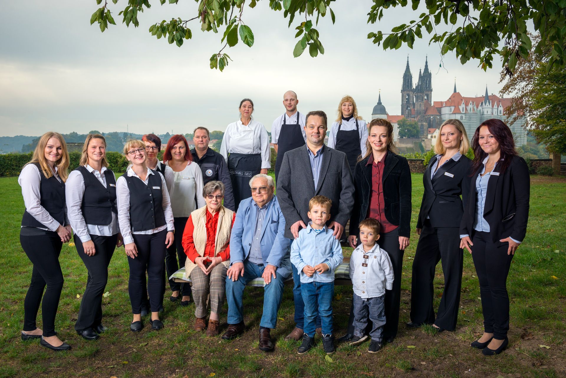 Eine Gruppe von Menschen posiert im Freien, einige in Uniformen, im Hintergrund ist eine Burg zu sehen.