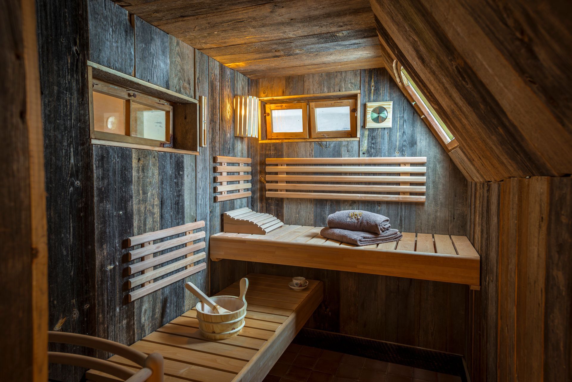 Rustikale Sauna aus Holz mit Bänken, Eimer und Handtüchern. Sonnenlicht strömt durch ein hohes Fenster herein.