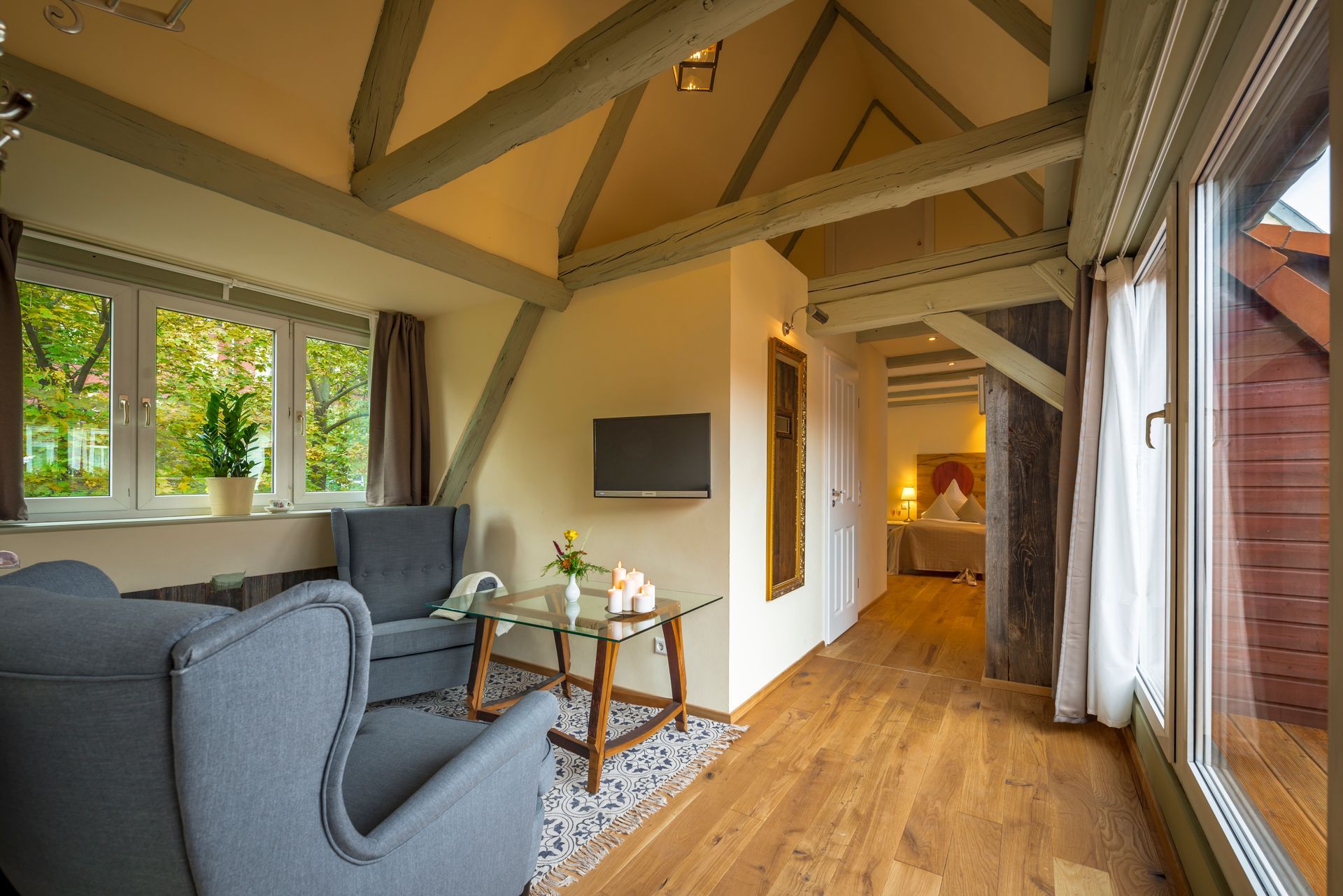Gemütliches Hotelzimmer mit Holzbalken, grauen Sesseln, einem kleinen Tisch, Fernseher und großen Fenstern mit Aussicht.