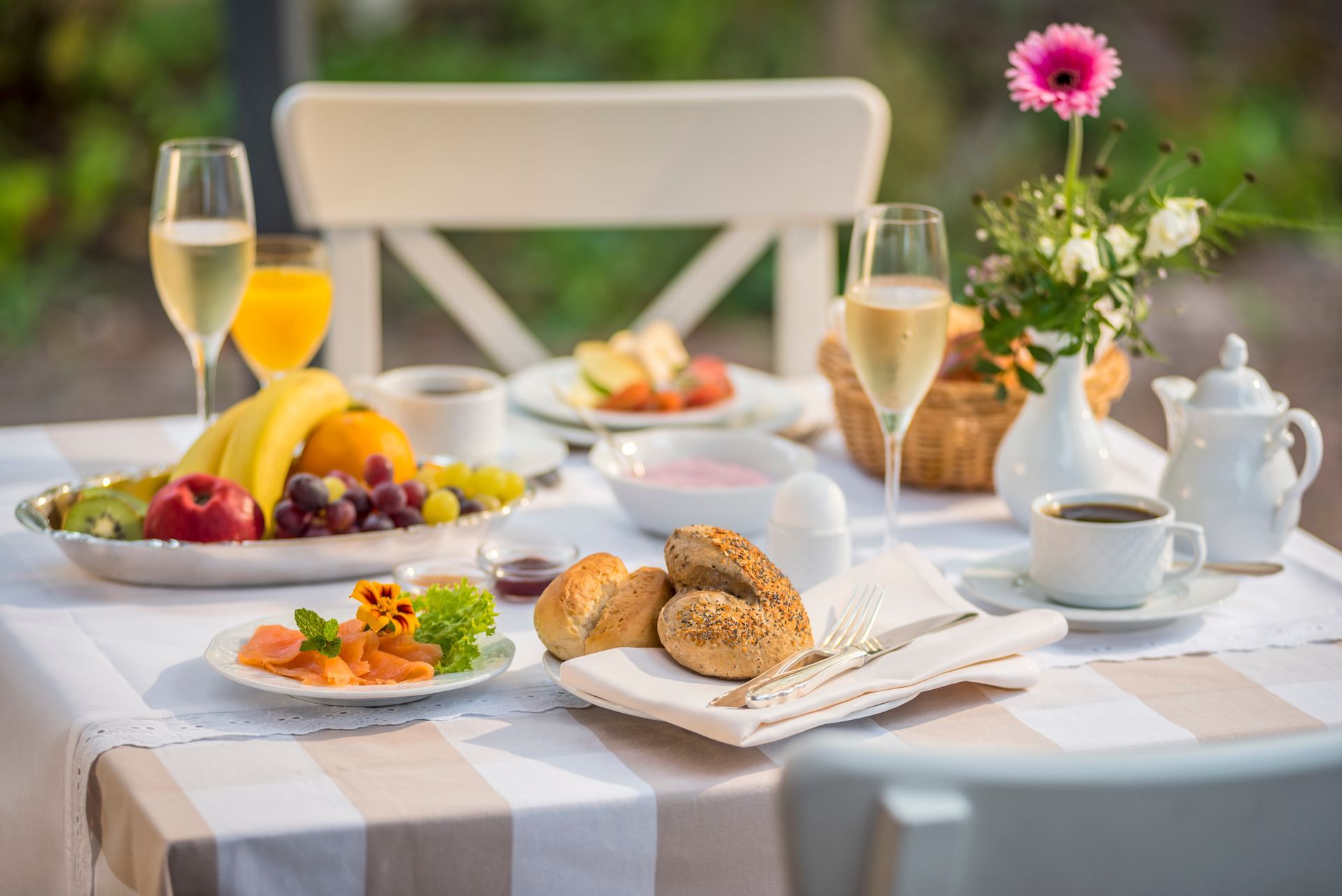 Ein Frühstückstisch im Freien ist gedeckt mit Obst, Brötchen, Kaffee und Champagner.