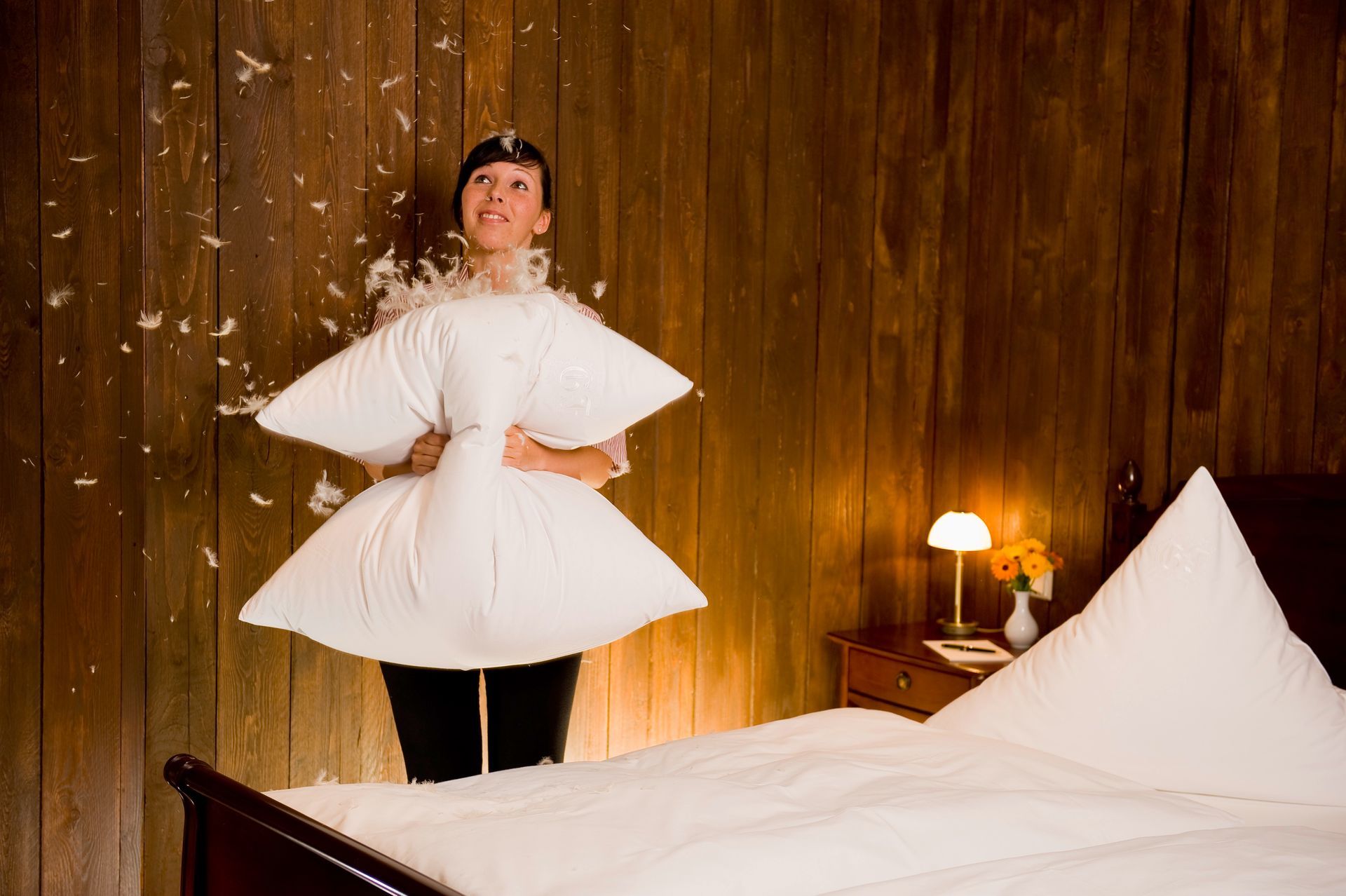 Frau im Bett, Kissen als Kostüm, Federn schweben. Holzwand und Nachttisch im Hintergrund.
