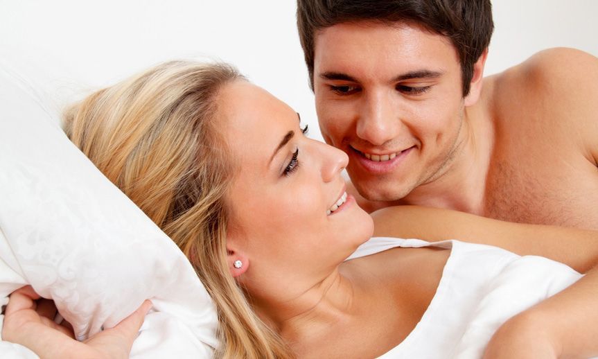 Ein Paar liegt einander zugewandt im Bett und tauscht liebevolle Lächeln vor einem schlichten weißen Hintergrund aus.