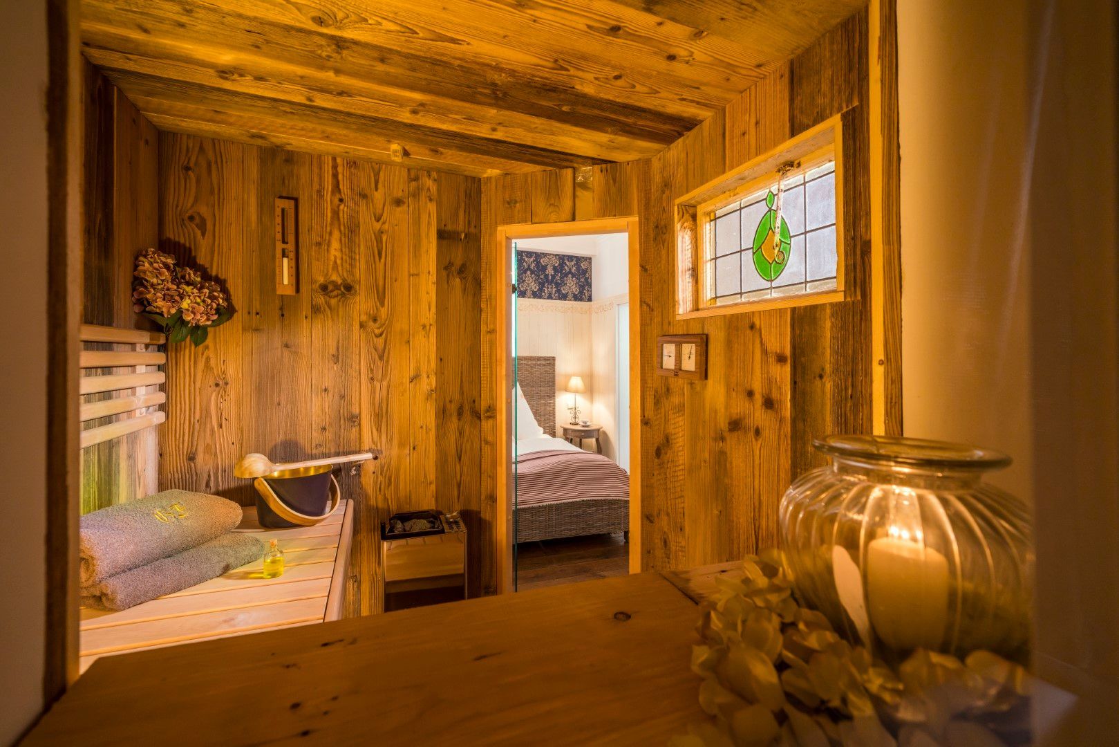 Innenansicht einer Sauna aus Holz, durch die Tür ist ein Bett sichtbar. Rechts befindet sich ein Fenster mit Buntglas und einem mit einer Kerze beleuchteten Gefäß.