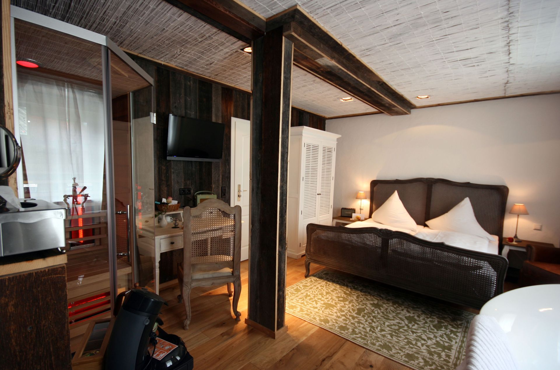 Schlafzimmer mit Holzbalken, dunklem Bett, Schreibtisch, weißem Kleiderschrank, Fernseher und Duschkabine aus Glas.