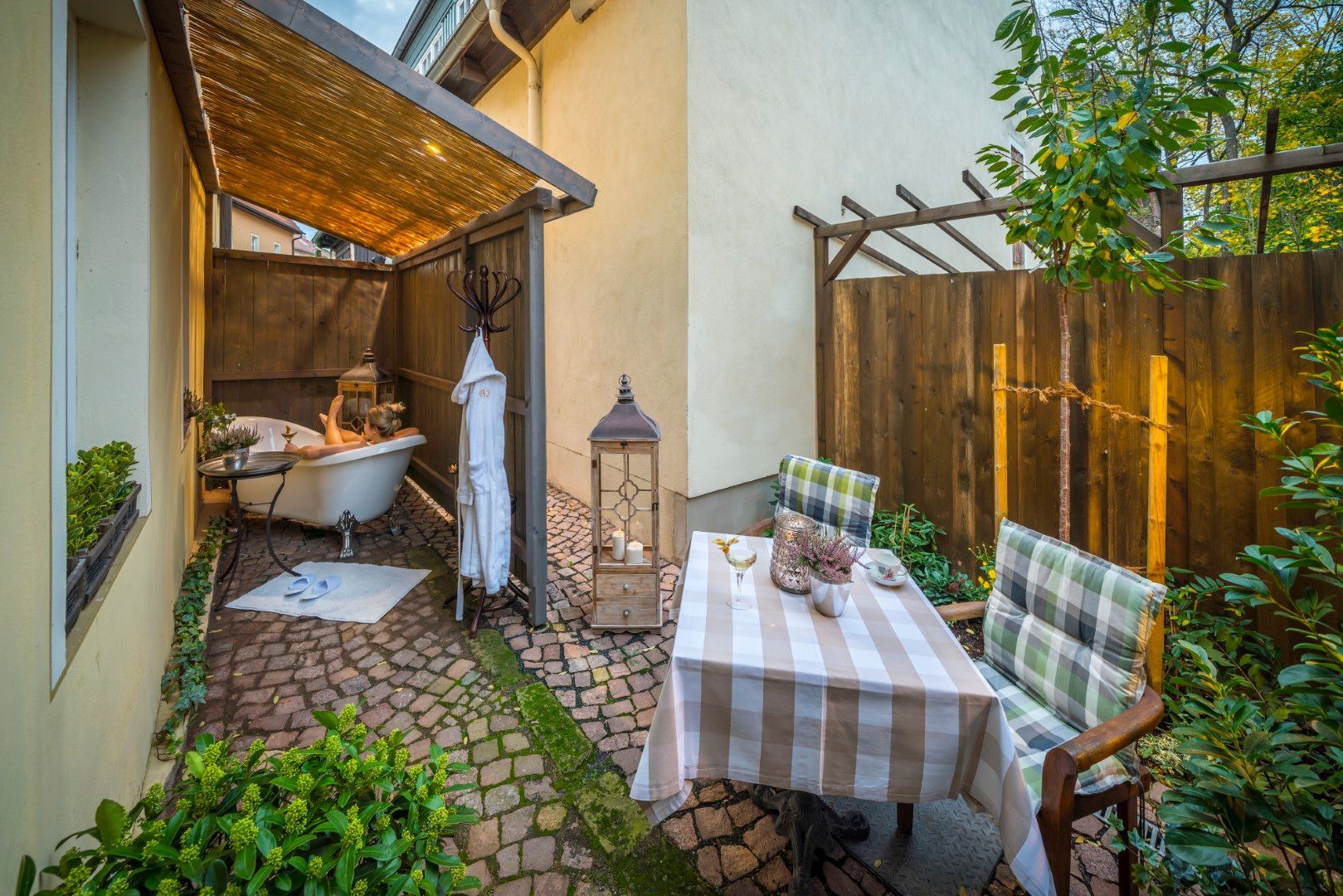 Outdoor-Spa: freistehende Badewanne, gestreifter Tisch, Stühle, Laterne und Pflanzen in einer gemütlichen Terrassenumgebung.