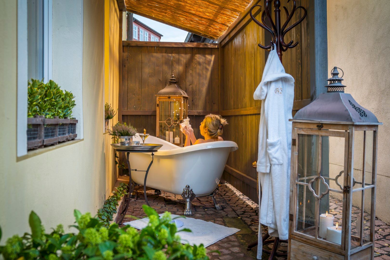 Frau entspannt in einer Badewanne im Freien, umgeben von Laternen und Grünpflanzen.