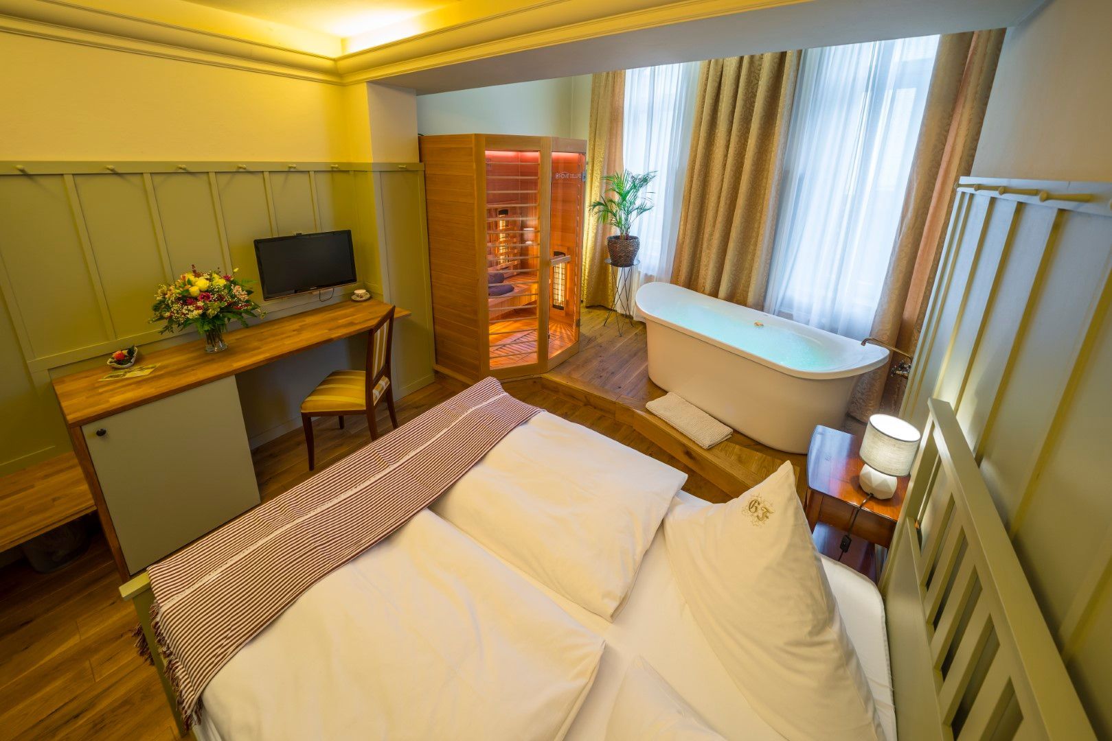 Schlafzimmer mit Bett, Schreibtisch und freistehender Badewanne; Sauna sichtbar. Beige- und Holztöne.