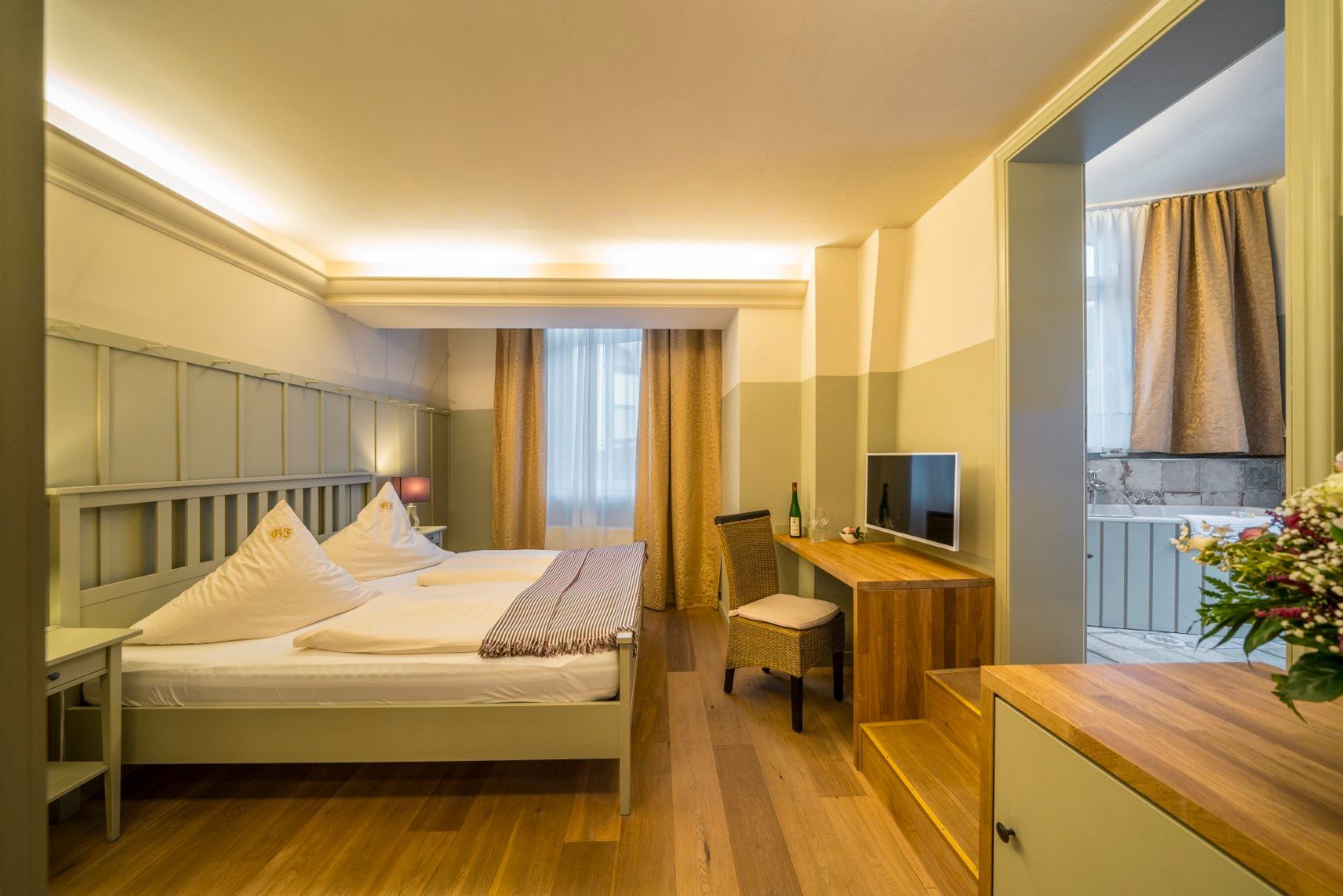 Hotelzimmer mit Bett, Schreibtisch, Fernseher und eigenem Badezimmer. Holzboden und neutrale Farbgestaltung.