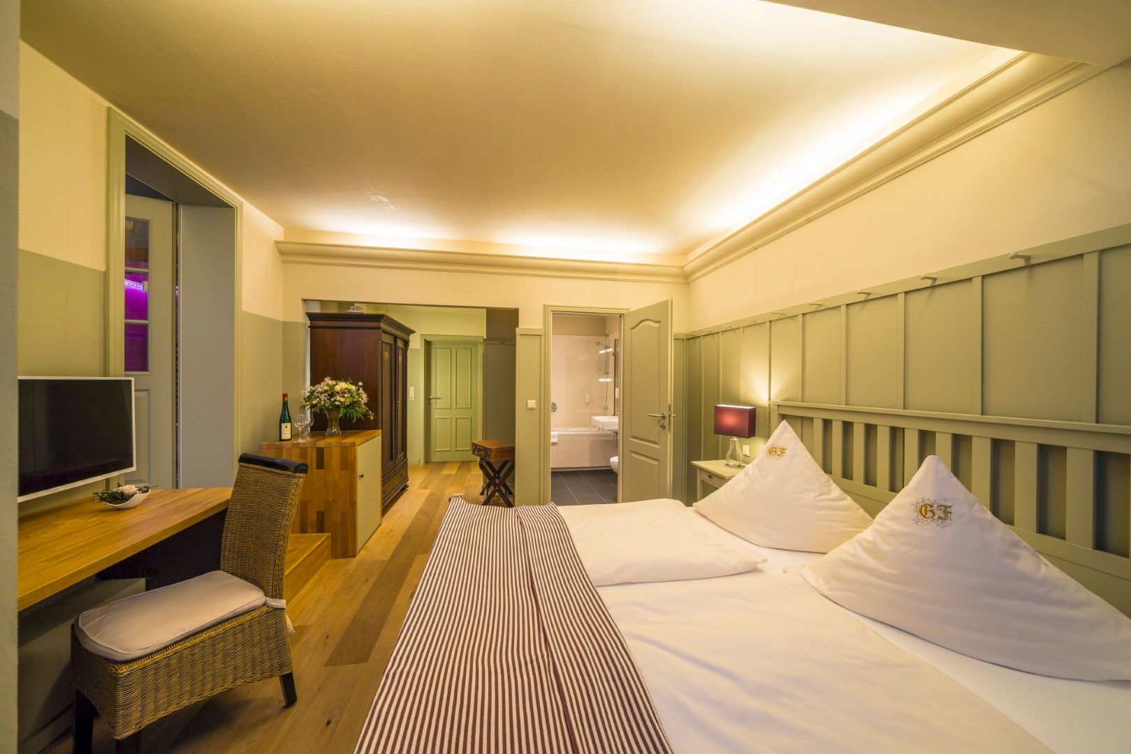Hotelzimmer mit Bett, Schreibtisch und eigenem Badezimmer. Holzböden und -vertäfelung, sanfte Beleuchtung.