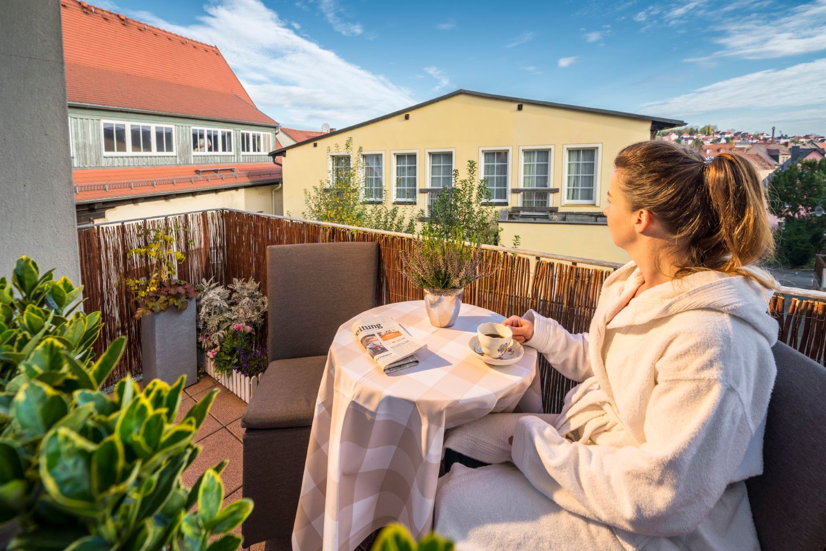 Eine Frau im Morgenmantel genießt ihren Kaffee auf einem Balkon mit Blick auf die Stadt.