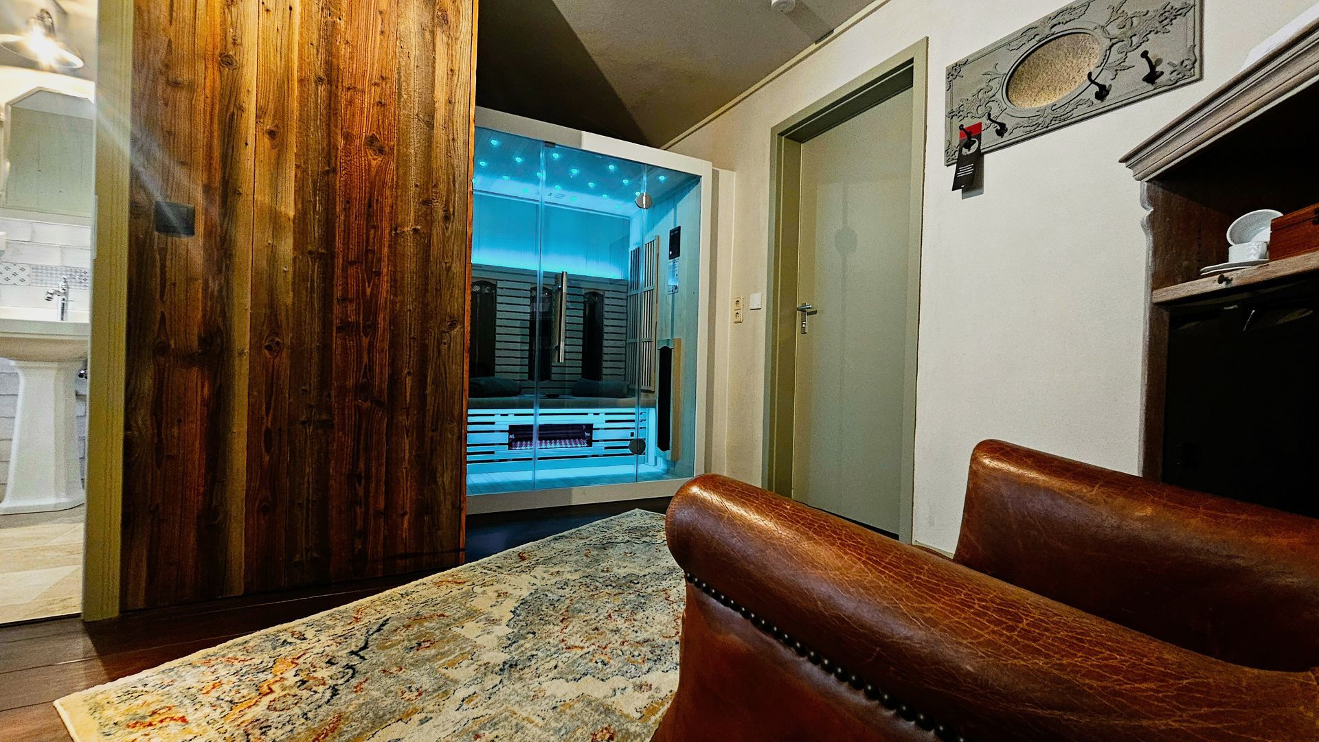 Die hölzerne Saunatür öffnet sich und gibt den Blick auf einen hellen Innenraum frei. Im Vordergrund sind Ledersessel und ein gemusterter Teppich zu sehen.