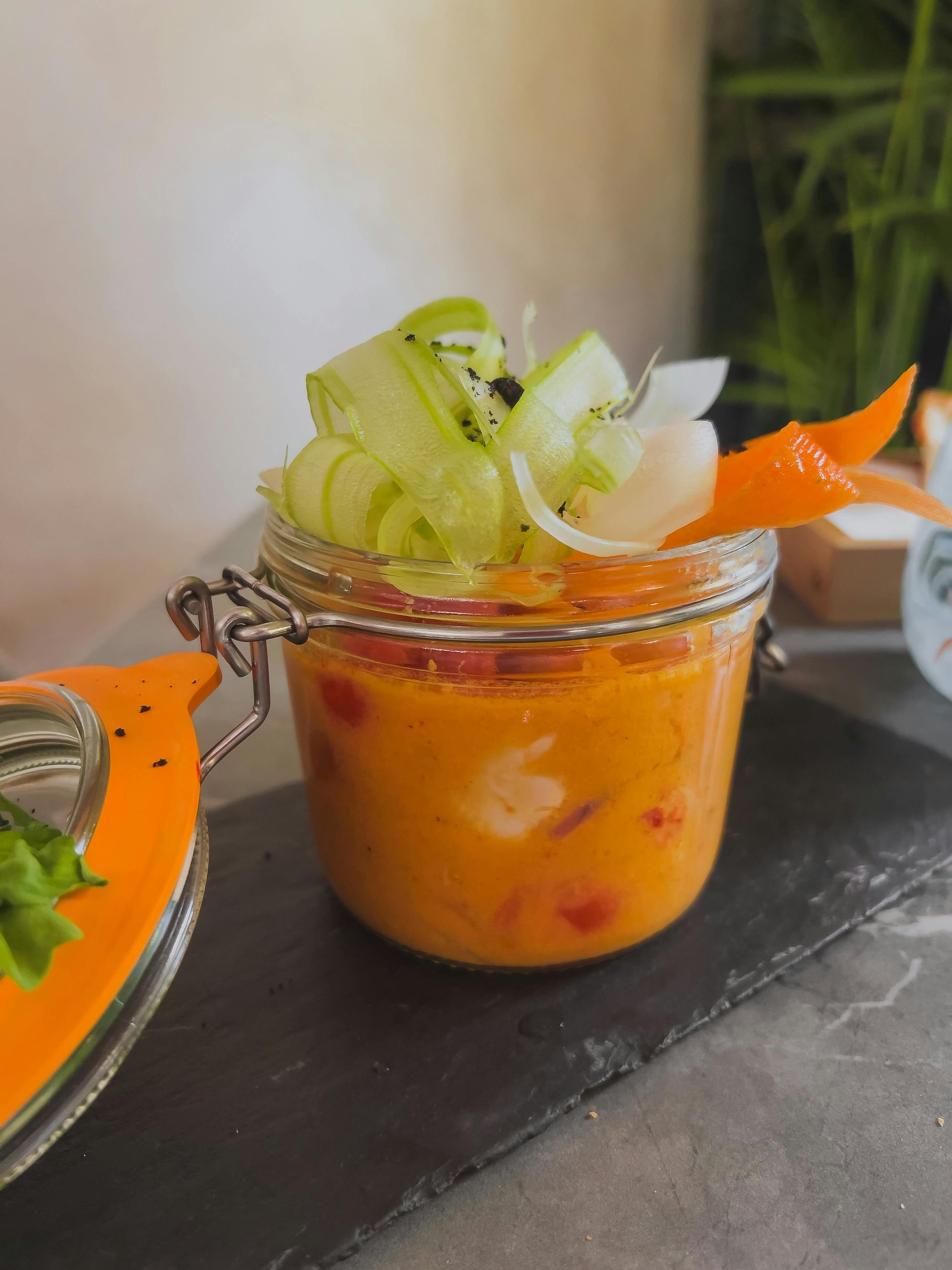 Ein mit Suppe gefülltes Glas, garniert mit geschnittenem Gemüse und einer orangefarbenen Karotte, auf einer schwarzen Schieferplatte, Nahaufnahme.