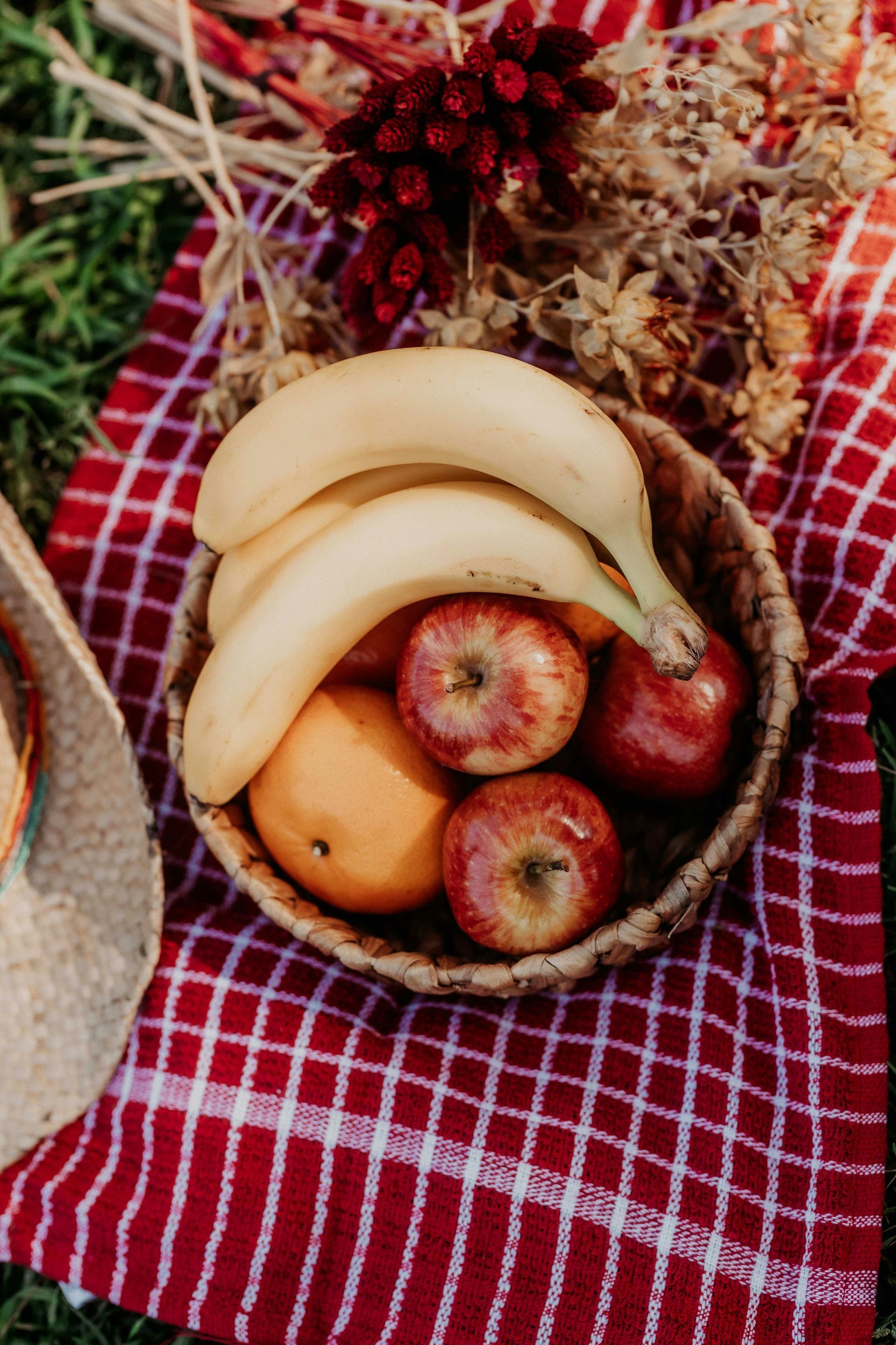 Ein geflochtener Korb, gefüllt mit zwei Bananen, zwei roten Äpfeln und einer Orange, steht auf einer rot-weiß karierten Picknickdecke.