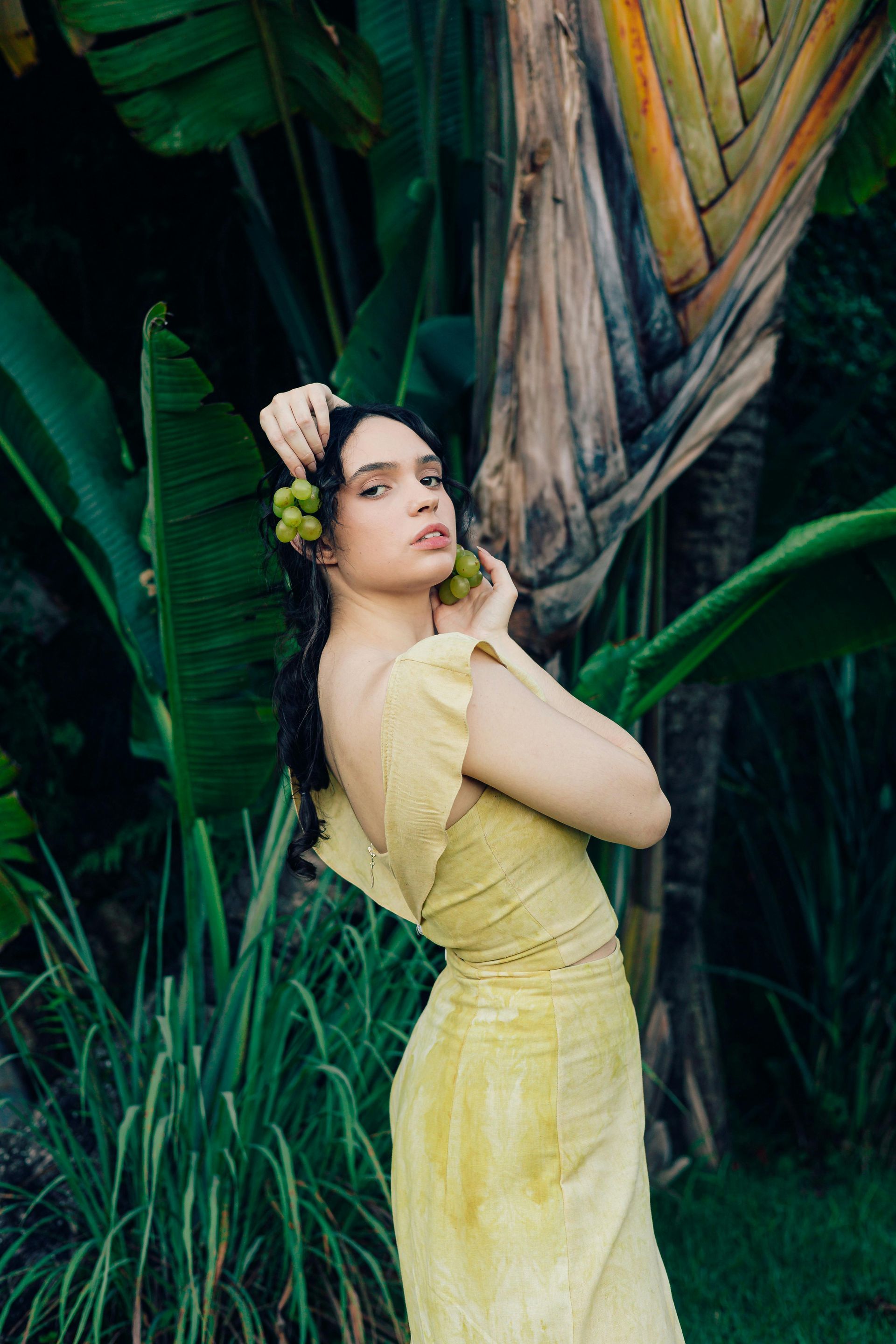 Frau in gelbem Kleid hält grüne Früchte; üppiges tropisches Laub im Hintergrund.
