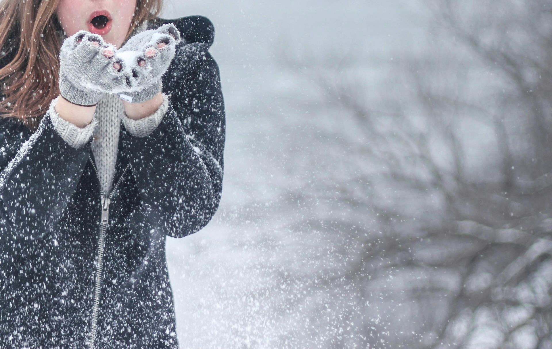 Eine Frau bläst in einer verschneiten Landschaft Schnee aus behandschuhten Händen.