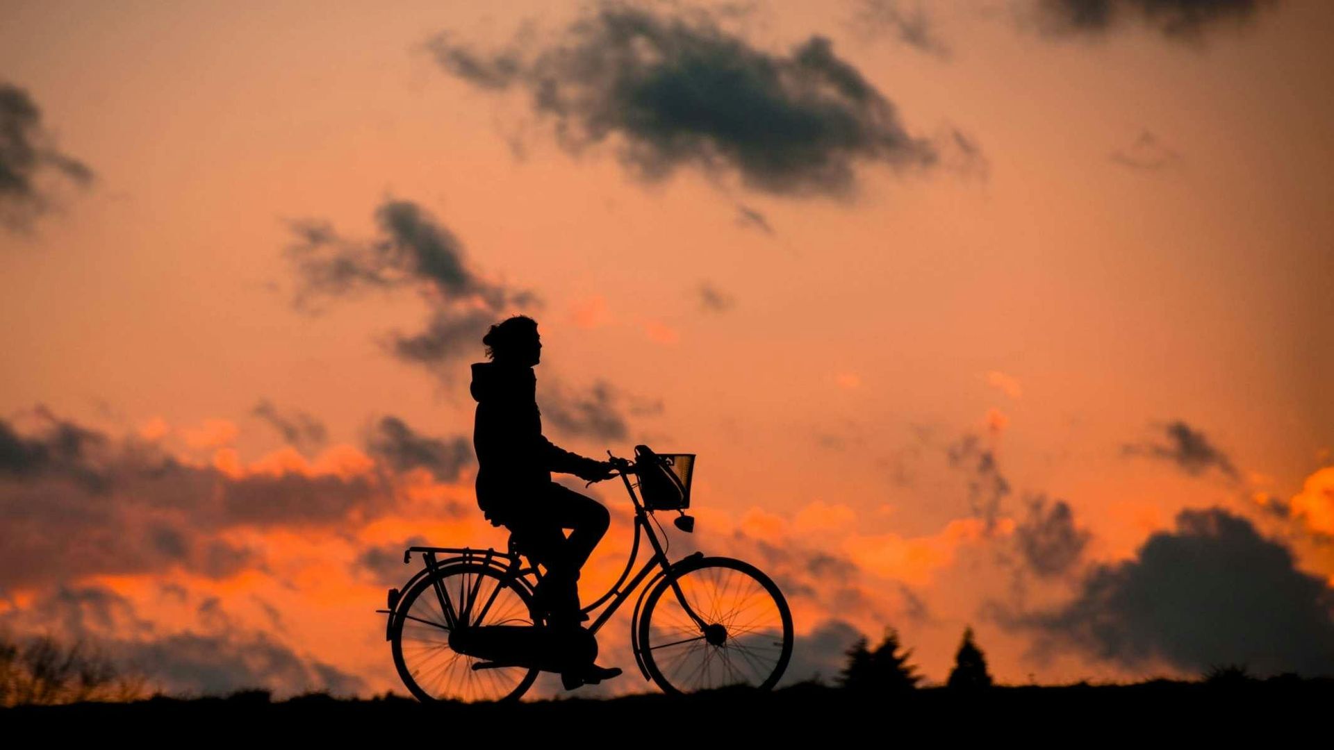 Die Silhouette einer Person, die auf einem Fahrrad fährt, vor einem leuchtend orangefarbenen Sonnenuntergangshimmel mit vereinzelten Wolken.