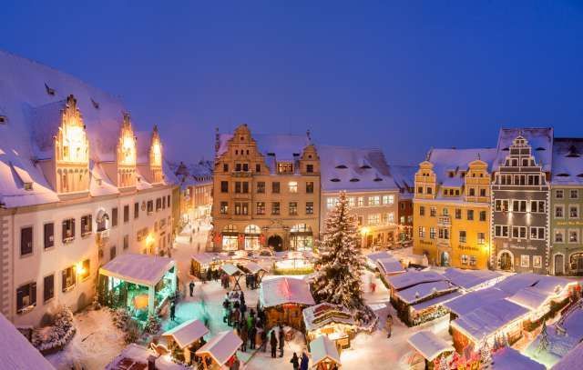 Ein verschneiter Weihnachtsmarkt auf einem europäischen Stadtplatz mit beleuchteten Ständen und Gebäuden unter dem Himmel der Abenddämmerung.