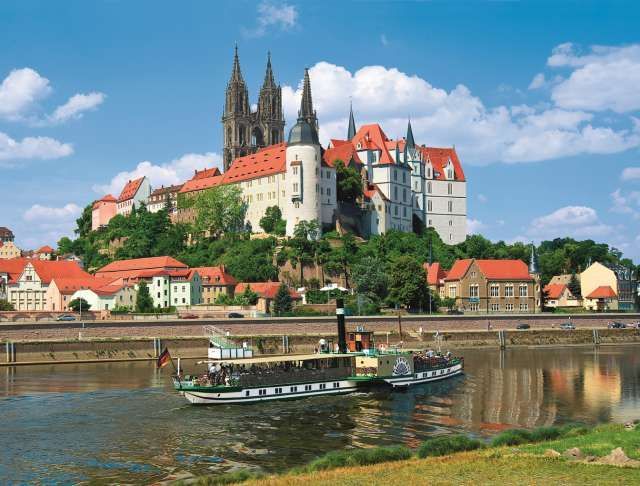 Malerische Ansicht einer Burg auf einem Hügel mit Blick auf einen Fluss, Boote, Gebäude und blauen Himmel.
