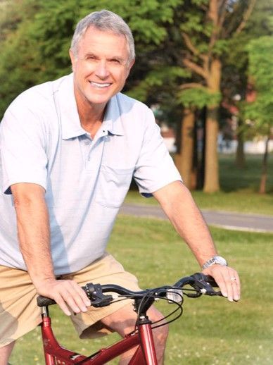 Ein Mann lächelt, während er auf einem roten Fahrrad durch einen Park fährt; er trägt ein hellblaues Hemd und hellbraune Shorts.