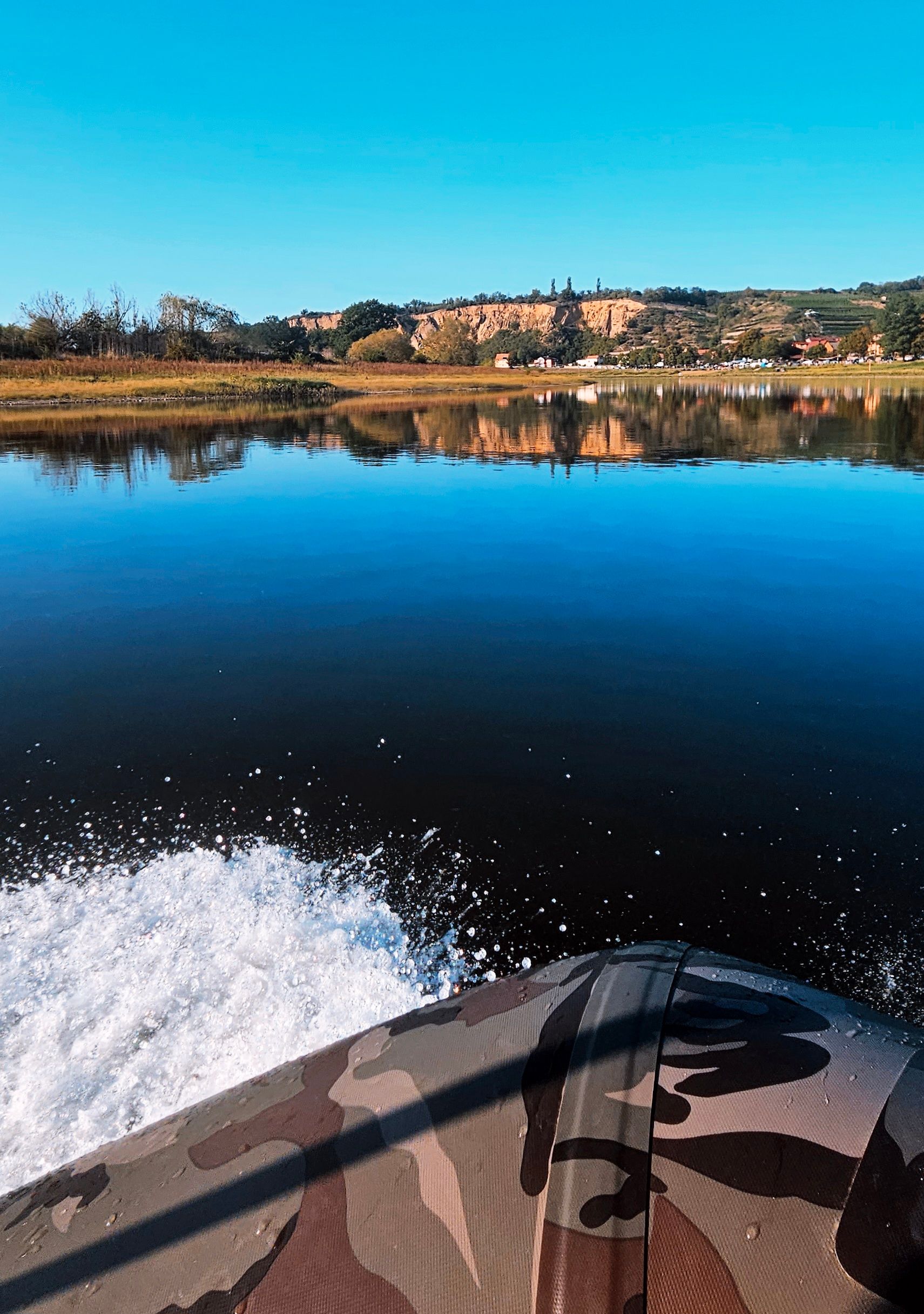 Boot auf dem Wasser, mit Tarnmuster, fährt auf ein von Bäumen gesäumtes Ufer und Gebäude zu, darüber strahlend blauer Himmel.