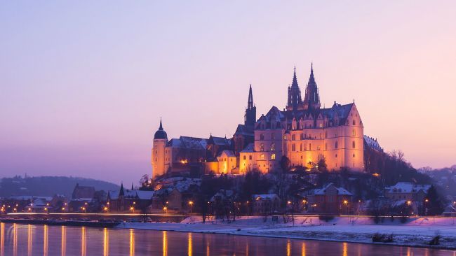 Beleuchtetes Schloss auf einem schneebedeckten Hügel, gespiegelt im ruhigen Wasser unter einem purpur-orangefarbenen Dämmerungshimmel.