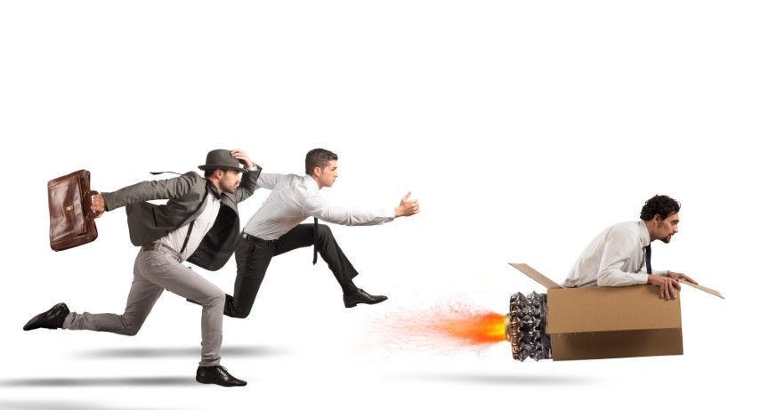 Drei Männer rennen: einer mit einer Aktentasche, ein anderer springt und der letzte sitzt in einer Pappkartonrakete mit Feuer.