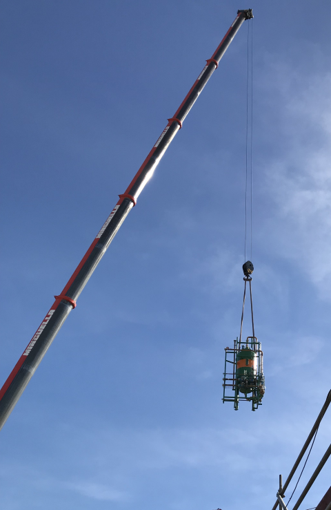 Kran hebt ein zylindrisches grünes Objekt in einem Käfig vor einem blauen Himmel.