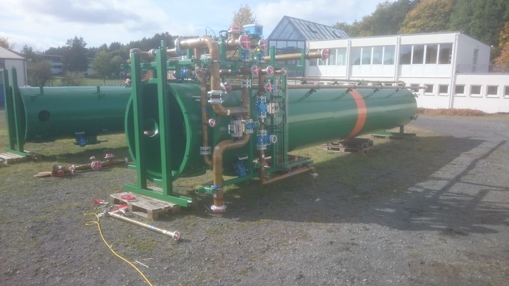 Grüne Industrietanks mit Rohrleitungen und Ventilen im Außenbereich vor einem Gebäude.