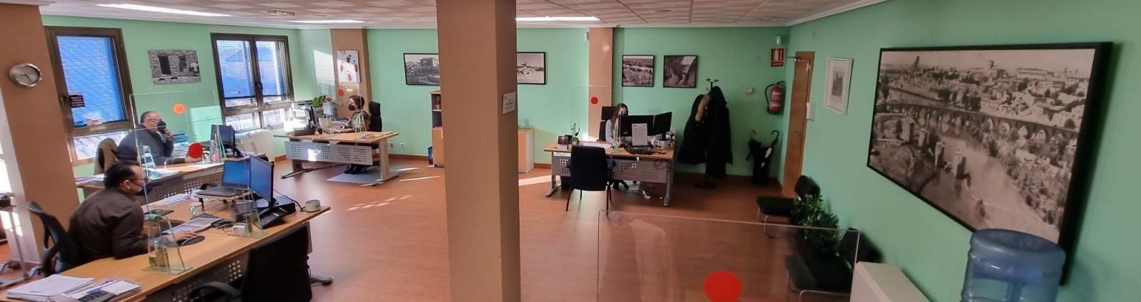 Una toma panorámica de una oficina luminosa con paredes verdes, escritorios de madera, personal trabajando en ordenadores y obras de arte en las paredes.