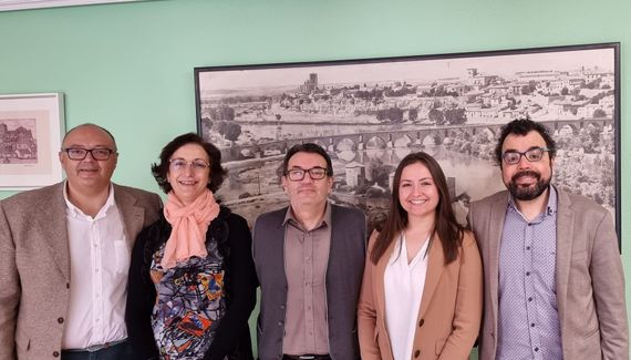 Cinco personas de pie, formando una fila, frente a una lámina enmarcada que representa una ciudad histórica, todas sonriendo a la cámara.