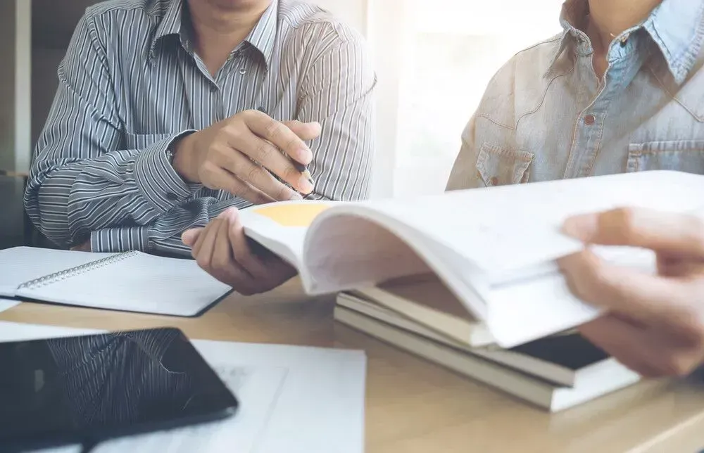 Personas revisando un libro abierto en una mesa con cuadernos y papeles cerca.