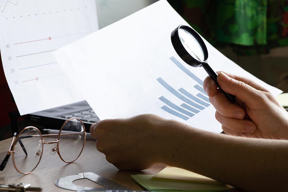Manos examinando gráficos financieros con una lupa junto a unas gafas sobre un escritorio.