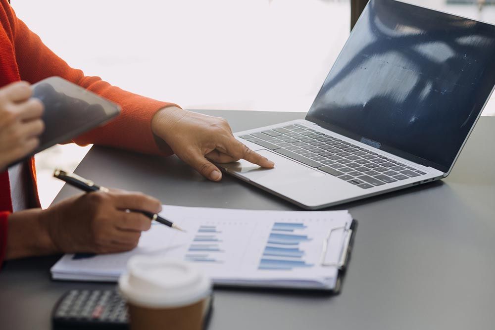 Dos personas revisando gráficos junto a una computadora portátil abierta sobre un escritorio con tazas de café.