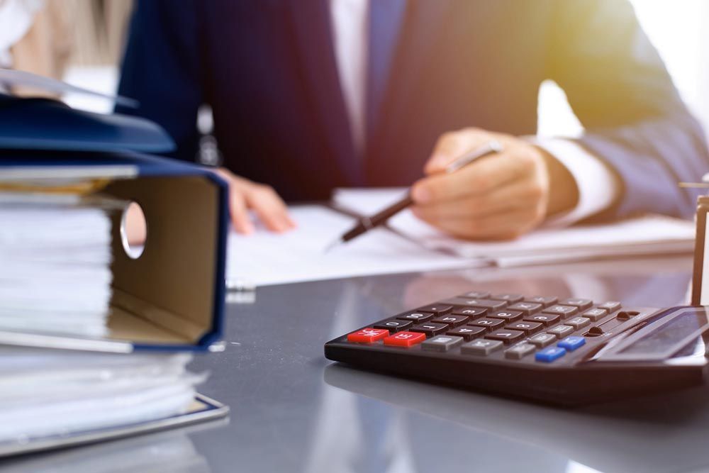 Una calculadora sobre el escritorio junto a una pila de carpetas mientras una persona toma notas al fondo.