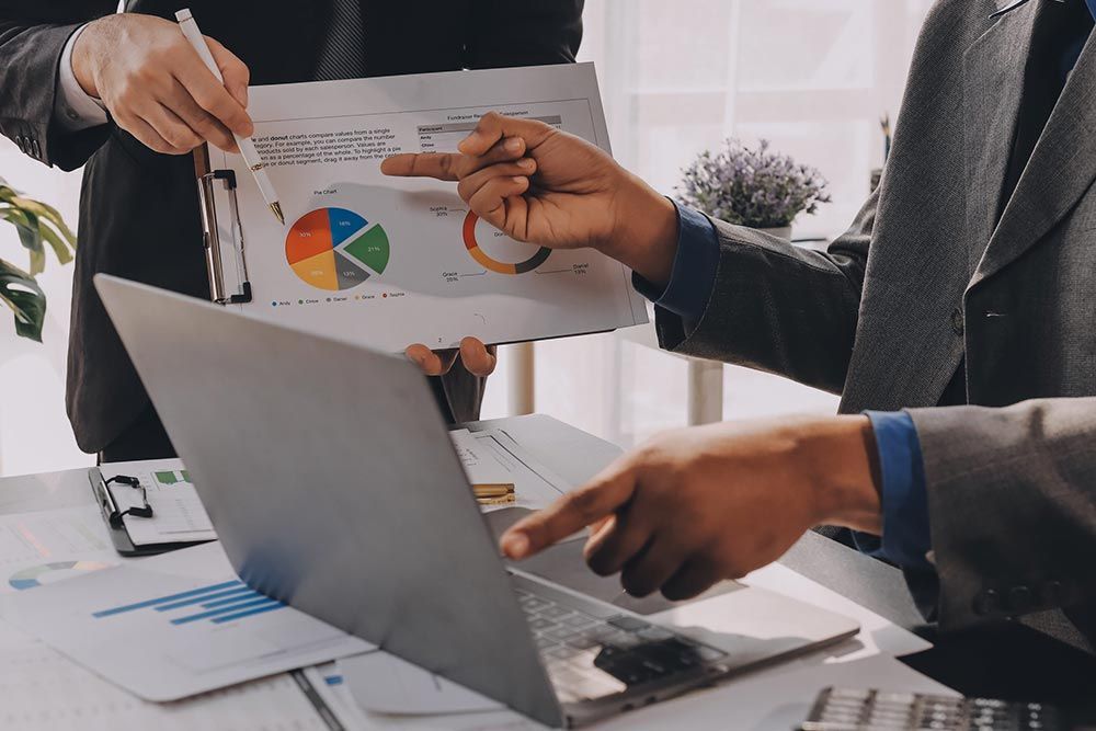 Compañeros revisando un gráfico circular colorido y documentos junto a una computadora portátil en una sala de reuniones.