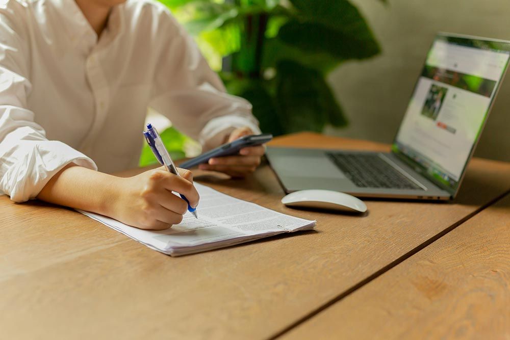 Una persona escribe en un cuaderno junto a un ordenador portátil sobre un escritorio de madera, con un ratón y una planta verde cerca.