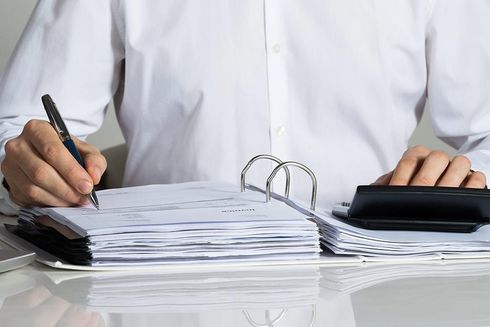Manos escribiendo en papeles junto a una carpeta y una calculadora sobre un escritorio.