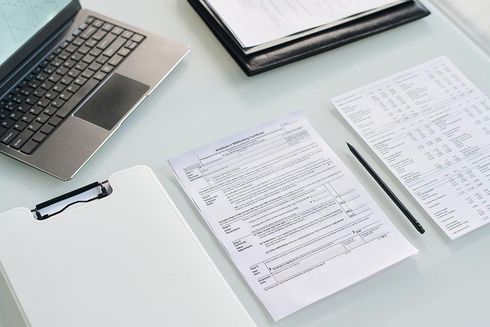 Vista superior de un ordenador portátil, un portapapeles, un bolígrafo y documentos financieros sobre un escritorio blanco.