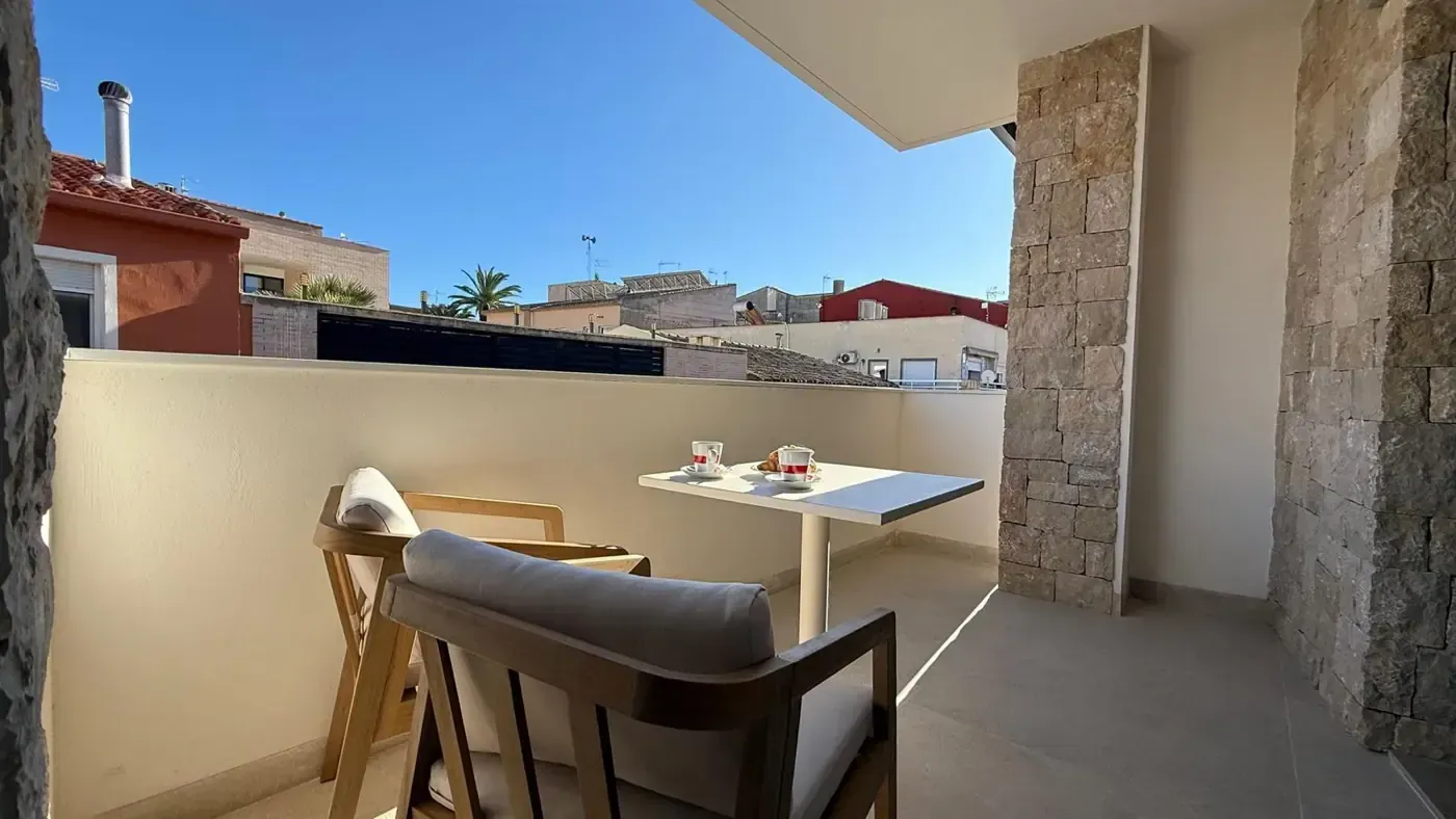 Un balcón soleado con dos sillas de madera con cojines color crema y una mesita puesta para desayunar bajo un cielo azul.